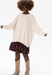 Kremowy, oversize sweter z szerokimi rękawami, w połączeniu z burgundową, warstwową spódnicą z koronkowymi akcentami oraz beżowymi kowbojkami.
