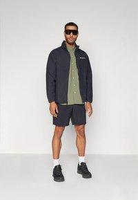 Uomo che indossa una giacca nera, una camicia verde oliva a bottoni, pantaloni corti neri, calzini bianchi, scarpe da ginnastica nere e grandi occhiali da sole scuri, in piedi su uno sfondo bianco.