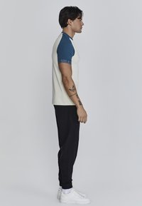 Korte mouwen raglan t-shirt in roomkleur en blauw, met blauwe accenten en tekst op de mouw, gecombineerd met zwarte joggingbroeken en witte sneakers.