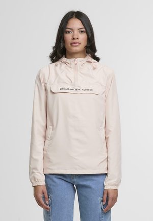 Young woman wearing a pale pink hooded jacket with "DREAM. BELIEVE. ACHIEVE." text, paired with light blue jeans, standing against white background.