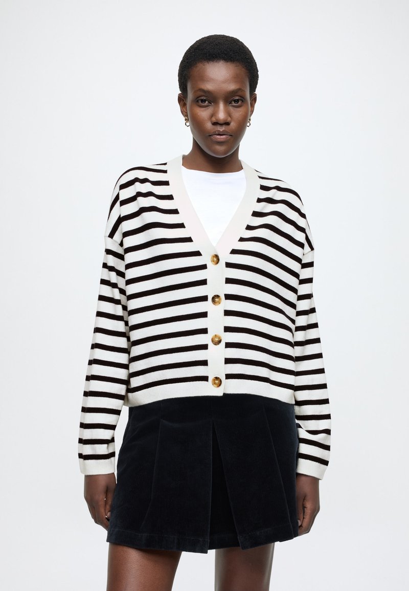 Svītrains melnas un krēmkrāsas kardigans ar V veida izgriezumu, zelta pogām un ribotām aprocēm. Savienots ar tumšu korduroja svārku ar krokām.