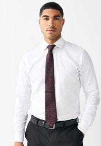 Weißes Hemd mit langen Ärmeln, dunkel gemusterte Krawatte mit Burgunder- und Schwarztönen, schwarzer Gürtel und dunkle Hose. Gepflegtes, maßgeschneidertes Erscheinungsbild.