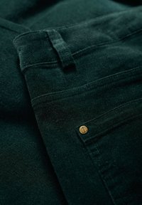 Ciemnozielona tkanina kordurowa o gładkiej fakturze, z złotym akcentem w kształcie kotwicy na kieszeni i przeszyciami.