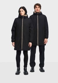 Svarta vadderade jackor med höga kragar och synliga dragkedjor. Båda modellerna bär svarta leggings och stövlar. Strukturert fleece-foder vid kragen.