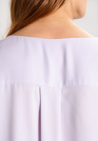Blusa de un suave color púrpura claro con una textura lisa, que presenta un escote ancho y pliegues en la parte posterior, destacando un diseño drapeado.