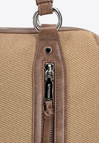 Detale torebki z brązowej teksturowanej tkaniny z skórzaną przywieszką suwaka, srebrnymi elementami i przeszywaną skórzaną ozdobą przy suwaku.