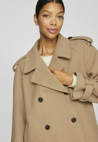 Beige dubbelknäppt kappa av mjukt tyg med stora slag, svarta knappar och axelremmar, lager på lager över en ljusfärgad tröja.