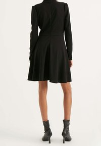 Robe noire à manches longues et corsage ajusté, jupe évasée et col montant. Tissu lisse et ourlet au genou. Bottines noires.