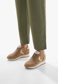 Brune ruskinds sneakers med en hvid gummisål. Funktioner inkluderer en polstret krave, snørebånd foran og et kontrasterende signaturlogo på siden.
