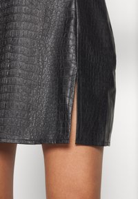 Gros plan d'une jupe noire en cuir synthétique avec une texture crocodile et une fente latérale laissant voir la peau en dessous.