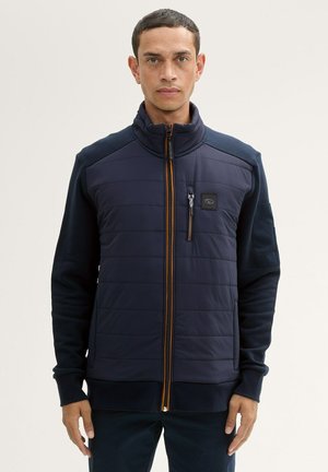 Navy padded jacket with high collar, quilted front, knitted sleeves, orange zipper detail, and chest pocket featuring a logo patch.