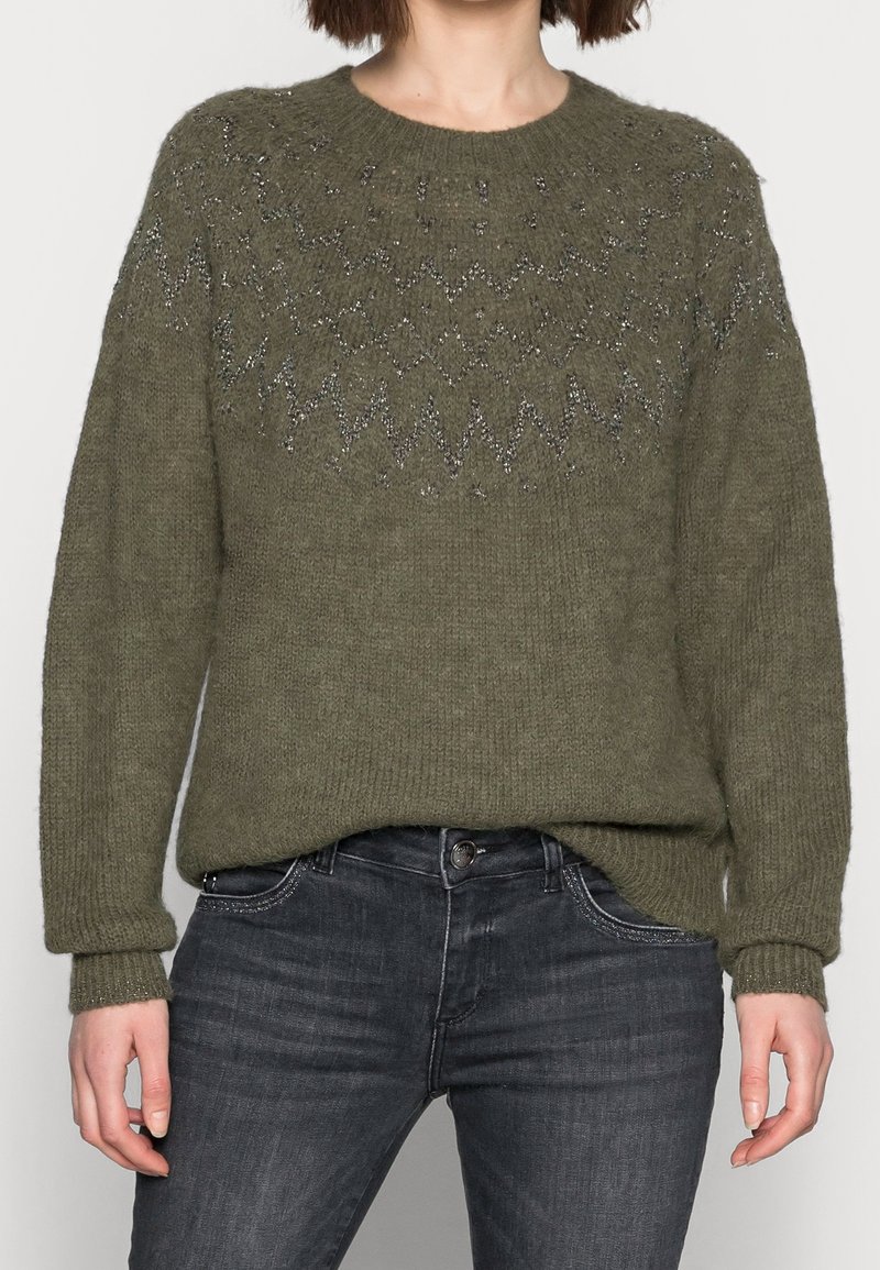 Femme portant un pull tricoté vert olive avec un motif zigzag métallique sur la poitrine et un jean gris foncé sur un fond uni.