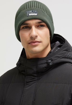 Green ribbed knit beanie with a branded patch. Worn by a person in a black puffer jacket with a high collar and drawstring adjustment.