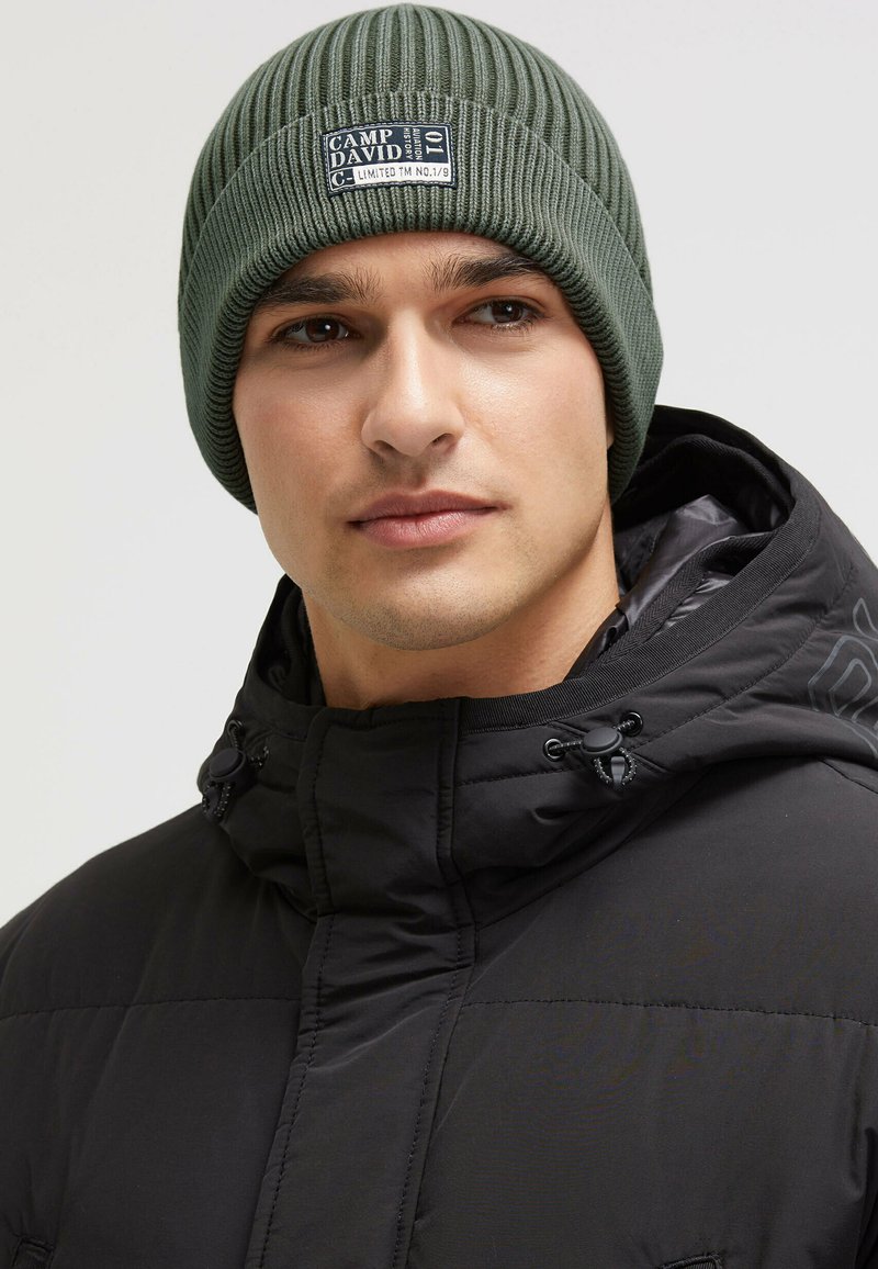 Green ribbed knit beanie with a branded patch. Worn by a person in a black puffer jacket with a high collar and drawstring adjustment.