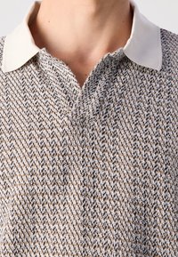 Gros plan d'une personne portant un haut en maille texturée et à motifs, avec un col tissé beige et un décolleté ouvert.