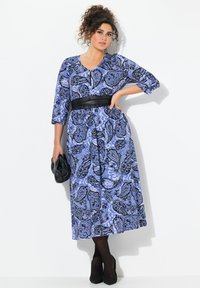 Vestido azul con estampado de paisley, de mangas tres cuartos, escote en V y cinturón negro en la cintura. Combinado con tacones negros y un bolso de mano negro.