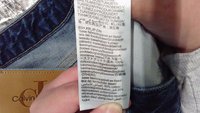 Étiquette indiquant la composition du tissu : 100 % coton ; 80 % coton, 20 % coton recyclé. Texte en plusieurs langues sur une étiquette en tissu blanc. Fond en denim.