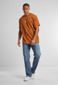 Bruine kortekorte mouwen t-shirt met een centrale witte print, gecombineerd met lichtblauwe jeans en witte sneakers. Effen kleuren, casual pasvorm.