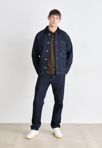 Denim jakna s džepovima, tamni traperice u istoj boji i bijele tenisice. Jakna ima metalne gumbe i teksturirani završetak.