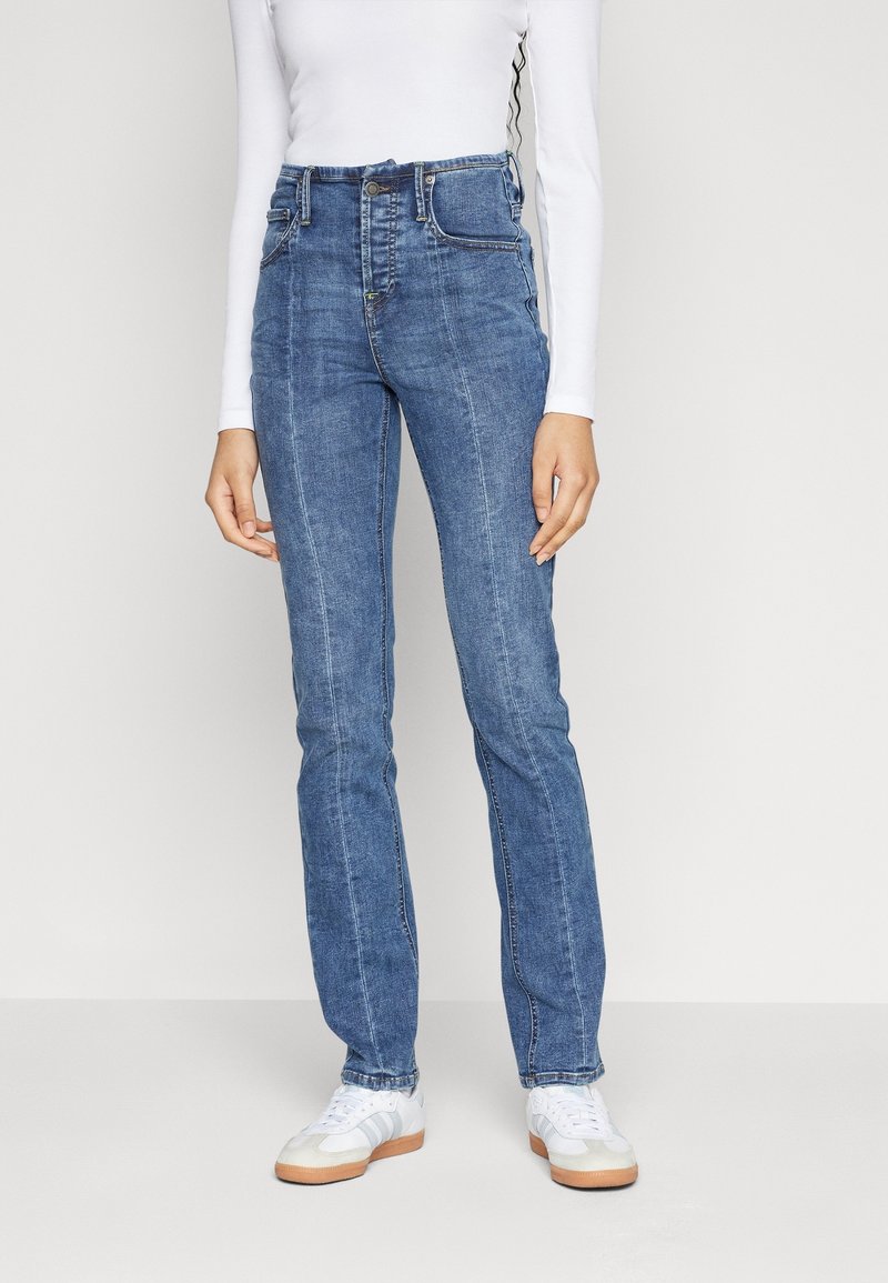 Jean en denim bleu taille haute avec une coupe droite, comportant cinq poches et des détails de couture visibles. Associé à des baskets blanches.