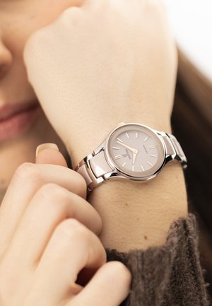 Ženska si prilagaja srebrno uro z belim številčnico in kovinsko zapestnico na levi roki, oblečena v rjavo jopico.