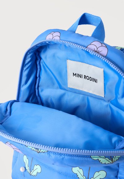 Sac à dos bleu pour enfants avec motif de fleurs violettes, partiellement ouvert montrant l'étiquette intérieure avec le texte "MINI RODINI".