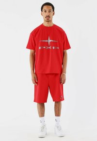 Rode T-shirt met korte mouwen en metallic "PXP" logo, gecombineerd met bijpassende shorts. Materiaal lijkt glad; relaxte pasvorm; gedragen met witte sneakers en sokken.