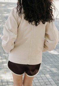 Veste oversized beige clair avec manches longues, associée à un short marron foncé avec un cordon blanc, sur un fond extérieur carrelé.