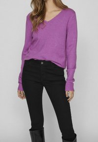 Pull violet à col en V en tissu tricoté doux, associé à un jean skinny noir. Le pull présente des manches longues avec des poignets côtelés.