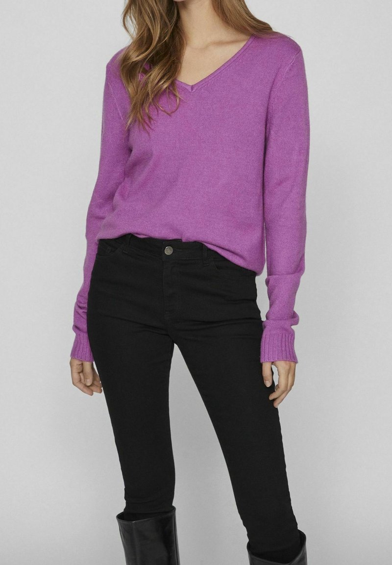 Pull violet à col en V en tissu tricoté doux, associé à un jean skinny noir. Le pull présente des manches longues avec des poignets côtelés.