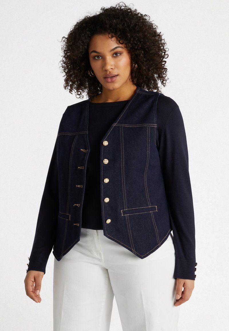 Marineblaues Denim-Weste mit goldenen Knöpfen, strukturierter Form und zwei Fronttaschen. Sie verfügt über einen V-Ausschnitt und einen schrägen Saum, kombiniert mit einem schwarzen Oberteil.