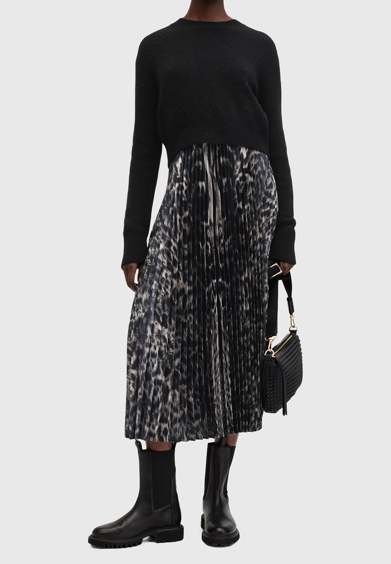Femme portant un pull noir, une jupe midi plissée à imprimé animal, des bottes en cuir noires et tenant un sac à main noir avec des accents dorés.