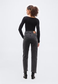 Γυναίκα με σγουρά μαλλιά στέκεται γυρισμένη με την πλάτη, φορώντας μαύρο μακρυμάνικο τοπ, ψηλόμεσα σκούρα γκρι τζιν και μαύρα μποτάκια σε λευκό φόντο.