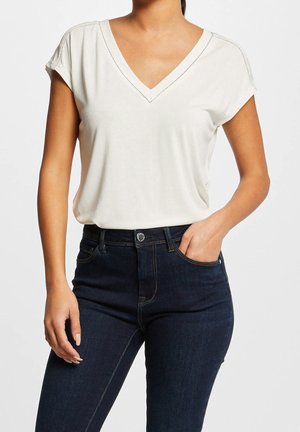 Maglietta a maniche corte color crema con scollo a V, vestibilità comoda e cuciture discrete sul collo, abbinata a jeans blu scuro con cinque tasche.