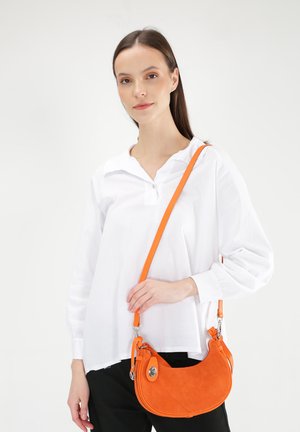 Oranje suède schoudertas met een gebogen vorm, zilveren hardware en een verstelbare schouderband, gecombineerd met een witte blouse en zwarte broek.