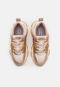 Baskets épaisses beige et or rose avec des lacets blancs, des accents métalliques et des détails pailletés, vues de dessus sur un fond blanc.