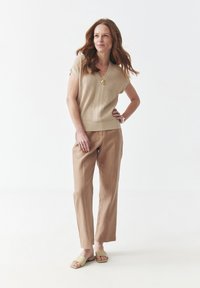 Top corto de punto beige con cuello en V y dobladillo acanalado, combinado con pantalones de lino marrón claro y sandalias deslizables color crema. Joyería minimalista como acento.