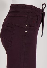 Close-up van donkerpaarse broeken met zijzakken, riemlussen en een strikkoord dat bij de taille is vastgebonden op een witte achtergrond.