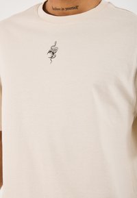 Beige Baumwoll-T-Shirt mit Rundhalsausschnitt, auf der Brust mit einem schwarzen Schlangenmotiv bestickt. Weicher Stoff, kurze Ärmel.