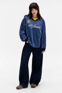 Camisa de mangas longas azul-marinho risca de giz com texto branco, combinada com jeans escuros de perna larga e sapatos cinzentos de slip-on. Ajuste casual.