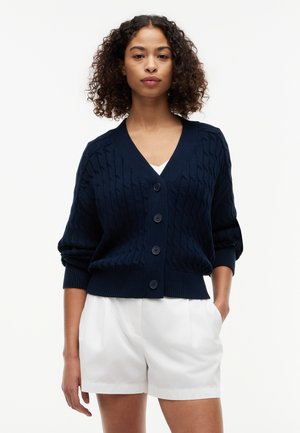 Žena s kučeravými vlasmi má na sebe tmavomodrý pletený zapínací sveter a biele šortky, stojí s jednou rukou v vrecku na jednoduchom pozadí.