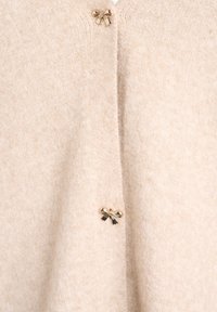 Bezs kašmira kardigāns, kas izgatavots no mīkstas vilnas, ar zelta krāsas pogām tuvojos lokā, kā akcentiem, un nedaudz teksturētu virsmu.