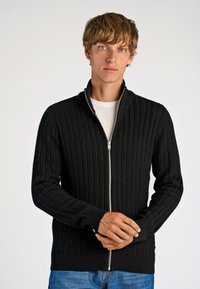Schwarzer Kabelstrick-Zipfel-Pullover mit hohem Kragen, gerippten Bündchen und Saum, kombiniert mit einem weißen Rundhals-Shirt und blauen Jeans.