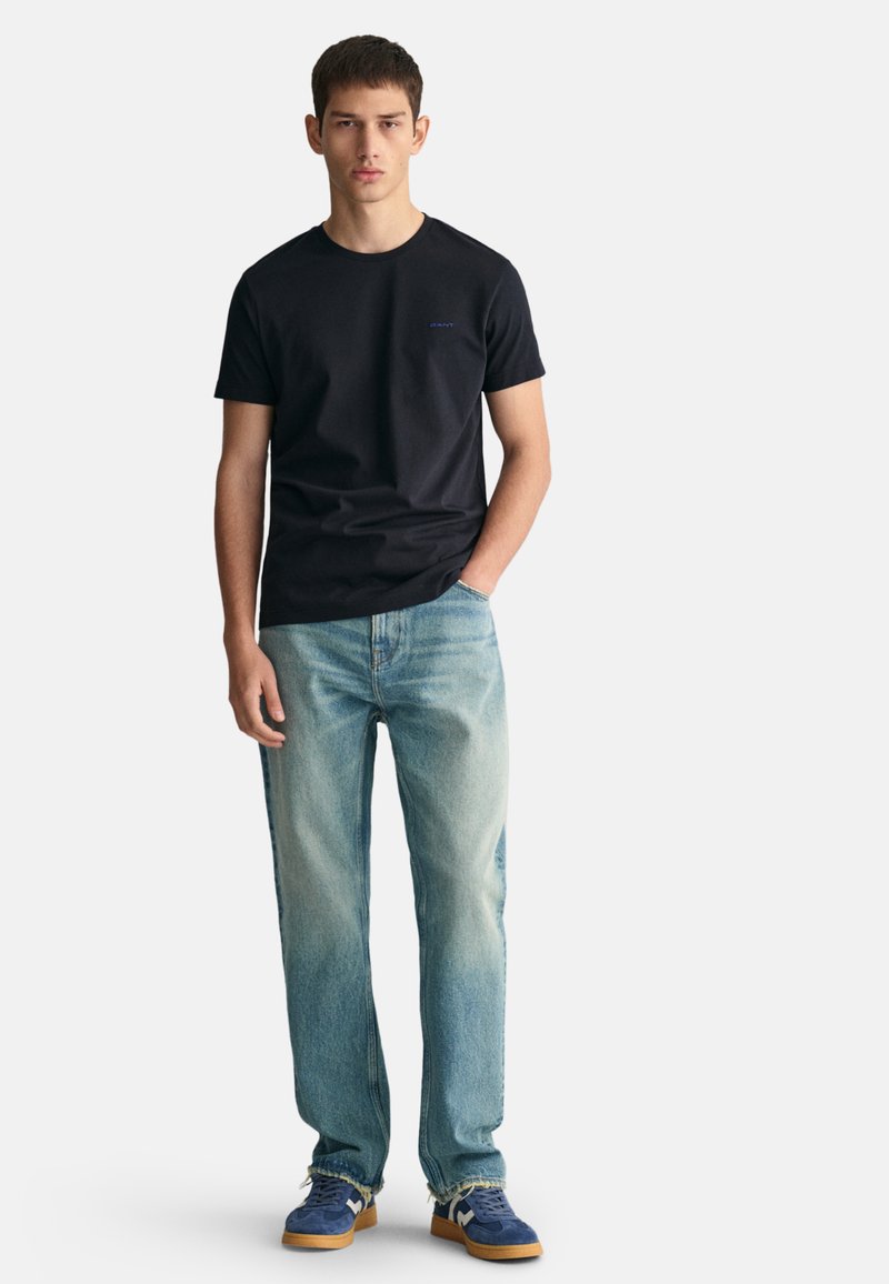 Camiseta de algodón negra con un pequeño logo azul, combinada con jeans de mezclilla azul claro y zapatillas azules con suelas beis, contra un fondo blanco.