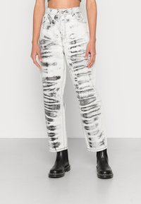Calças de ganga brancas com padrão tie-dye preto, de cintura alta, perna direita e costura visível. Usadas com botins pretos.