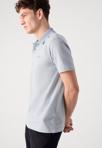 Lys blå polo skjorte med en to-knappers placket, korte ermer, hvite krageakenter og en tonalt logo på brystet. Glatt stofftekstur.