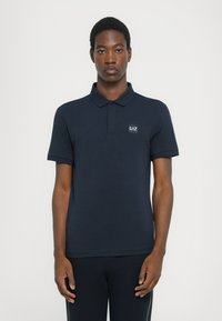 Camisola polo azul marinho feita de algodão. Apresenta um decote com gola e mangas curtas, com um pequeno emblema da marca no lado esquerdo do peito.