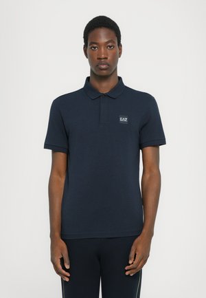 Marineblå polo-shirt laget av bomull. Har en krage og korte ermer, med et lite merketempel på venstre bryst.