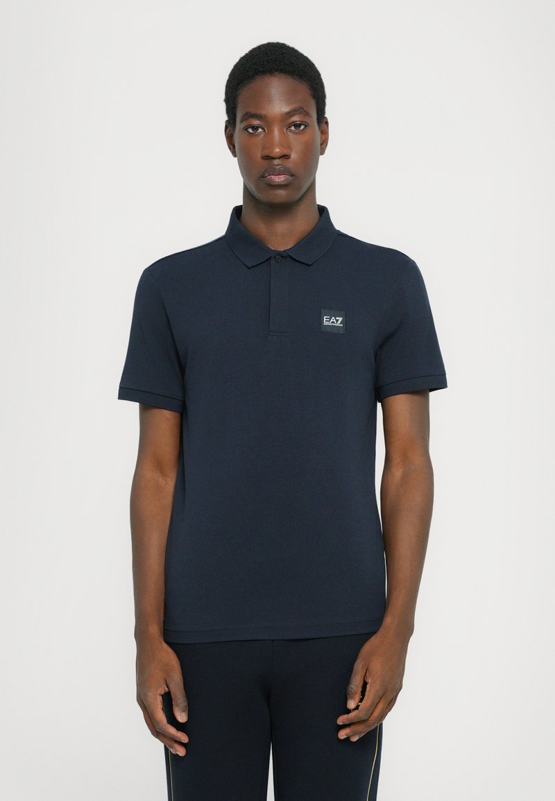 Camisola polo azul marinho feita de algodão. Apresenta um decote com gola e mangas curtas, com um pequeno emblema da marca no lado esquerdo do peito.