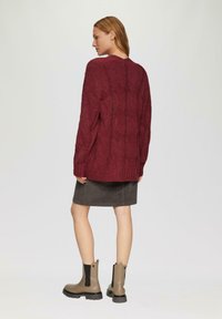 Bordeauxfarbener Strickpullover mit langen Ärmeln, strukturiertem Muster und gerippten Bündchen. Kombiniert mit einem grauen Rock und taupefarbenen Stiefeletten.